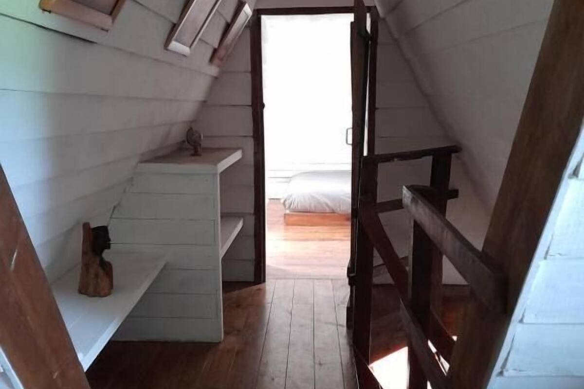 Un dormitorio bajo mansárdico con paredes y piso de madera, un pequeño tocador blanco y una cama con colchón blanco.