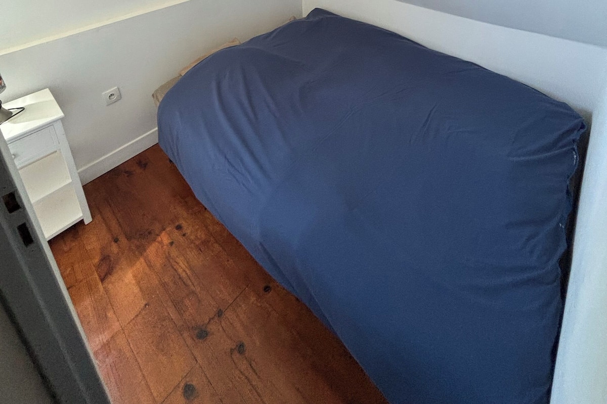 Una habitación pequeña con cama individual, suelo de madera, mesita de noche blanca y colchón azul.