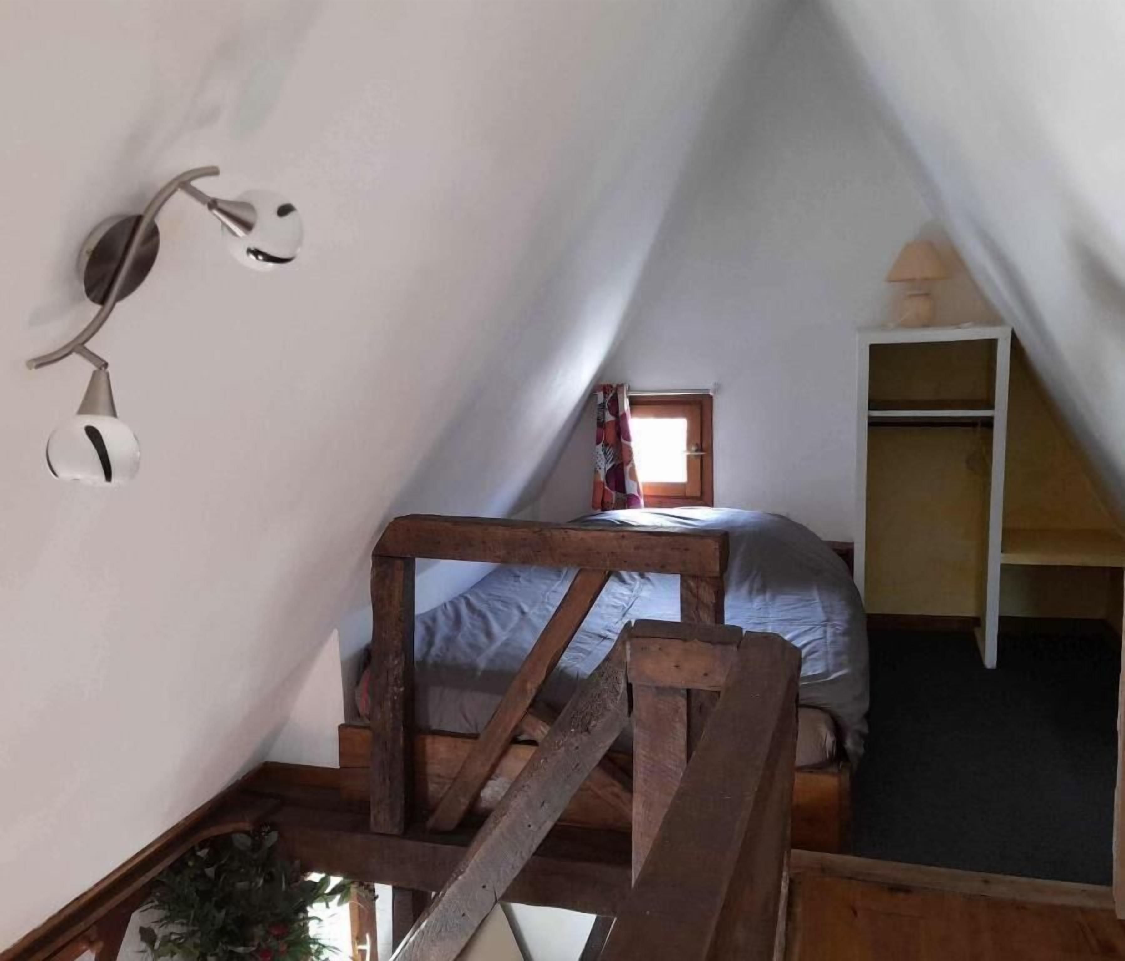 A small attic bedroom with a wooden bunk bed, a small window with curtains, a wall-mounted lamp, and a wooden ladder.