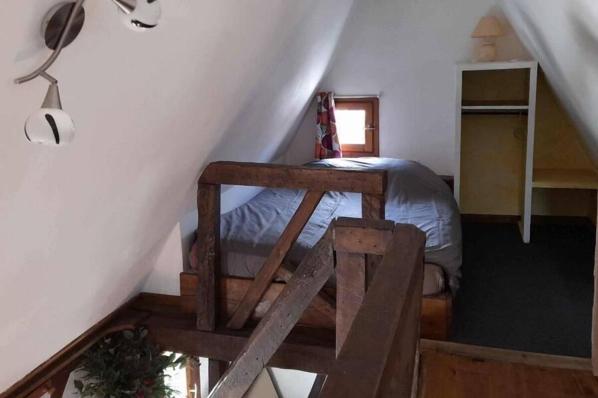 A small attic bedroom with a wooden bunk bed, a small window with curtains, a wall-mounted lamp, and a wooden ladder.