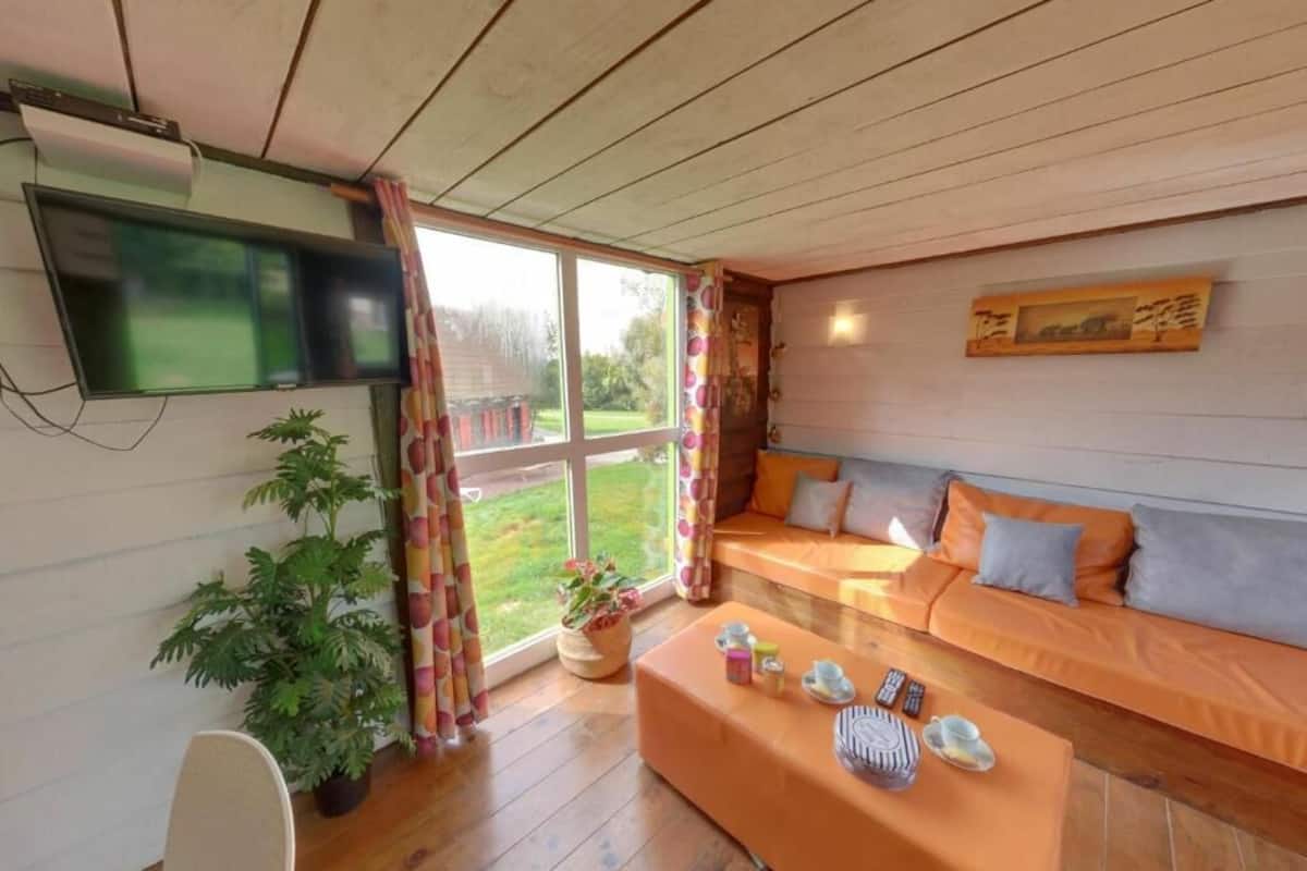 Una sala de estar acogedora con piso de madera, un conjunto de sofá, un televisor de pantalla plana y un ventanal con cortinas.