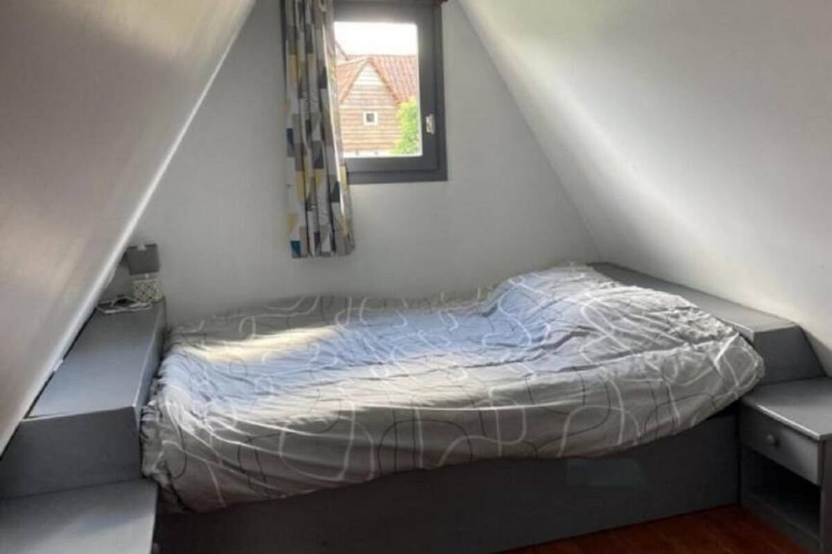 Habitación pequeña con techo inclinado, una cama, un ventanal y vista a una casa.