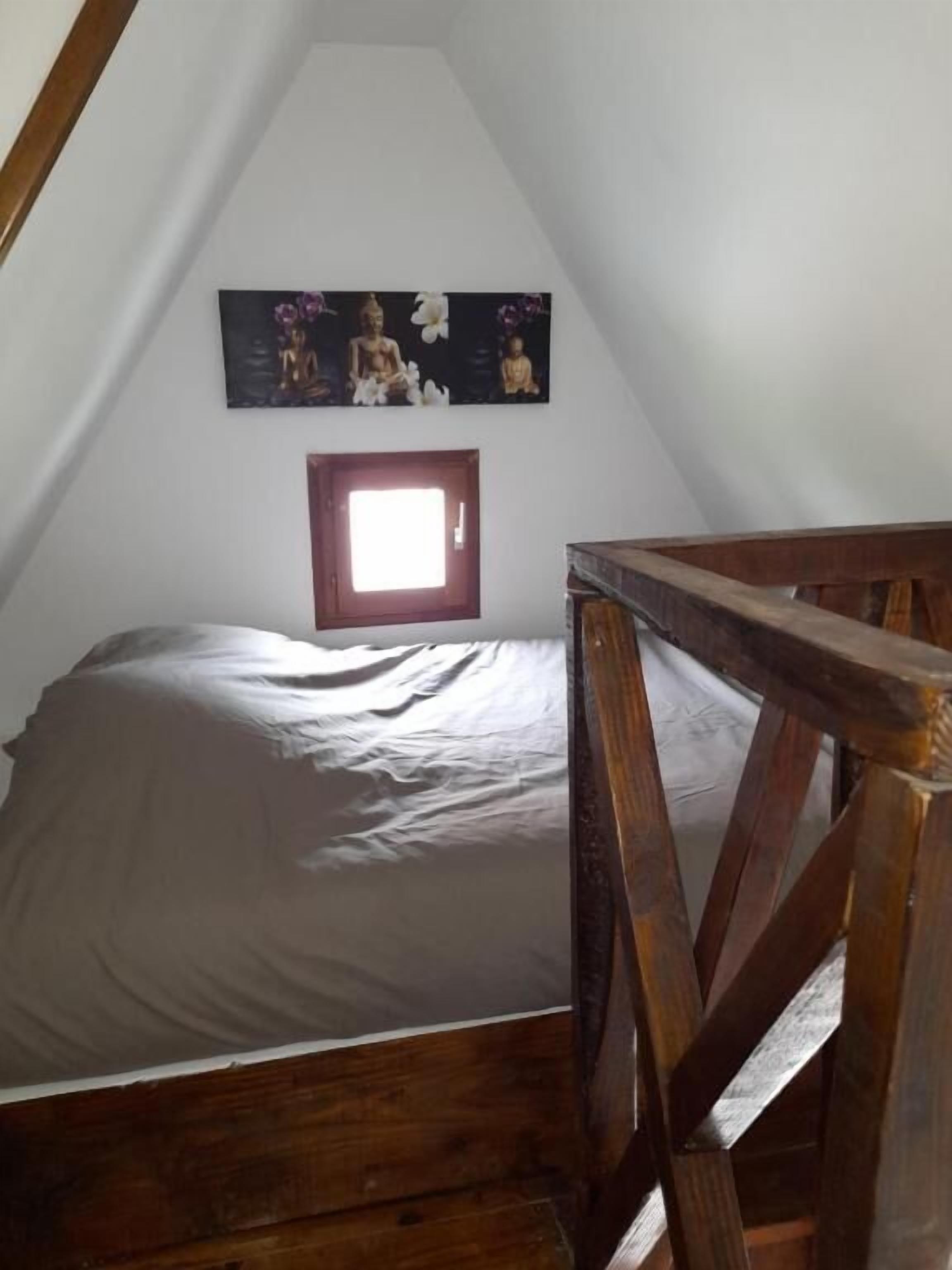 A bedroom with a sloped ceiling, a bed with a gray sheet, a wooden headboard, and a wall-mounted picture of Buddha statues.