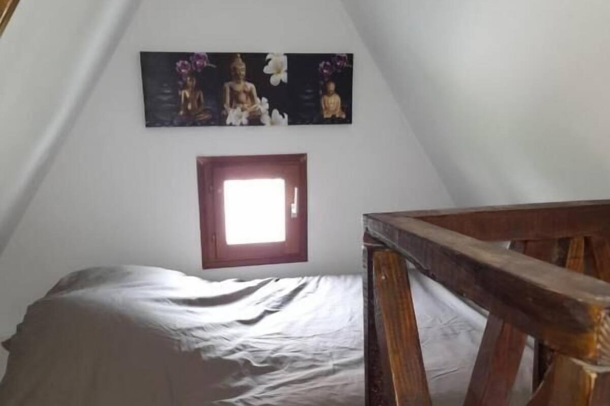 Un dormitorio con techo inclinado, cama con sábana gris, cabecero de madera y un cuadro en la pared con estatuas de Buda.