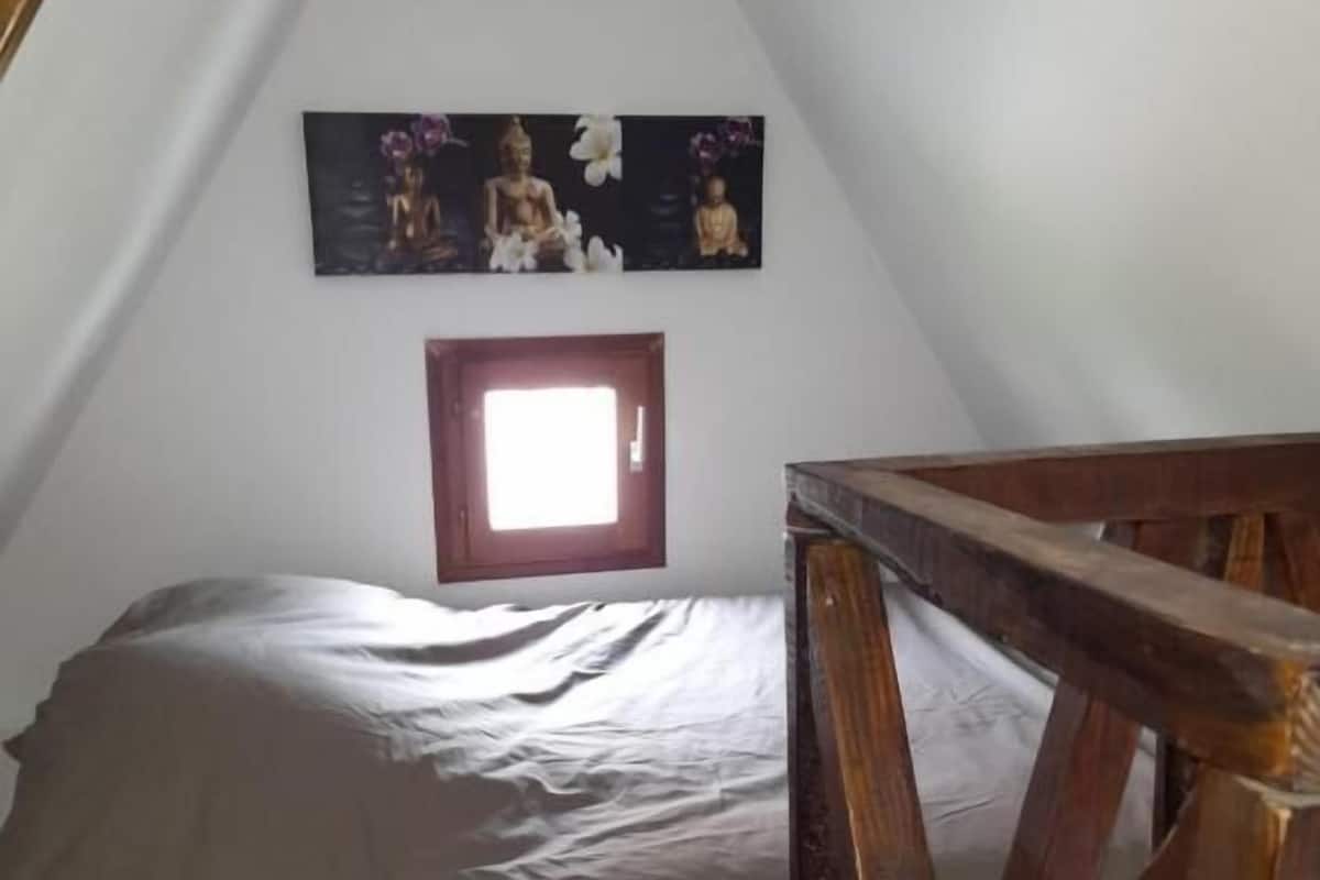 A bedroom with a sloped ceiling, a bed with a gray sheet, a wooden headboard, and a wall-mounted picture of Buddha statues.