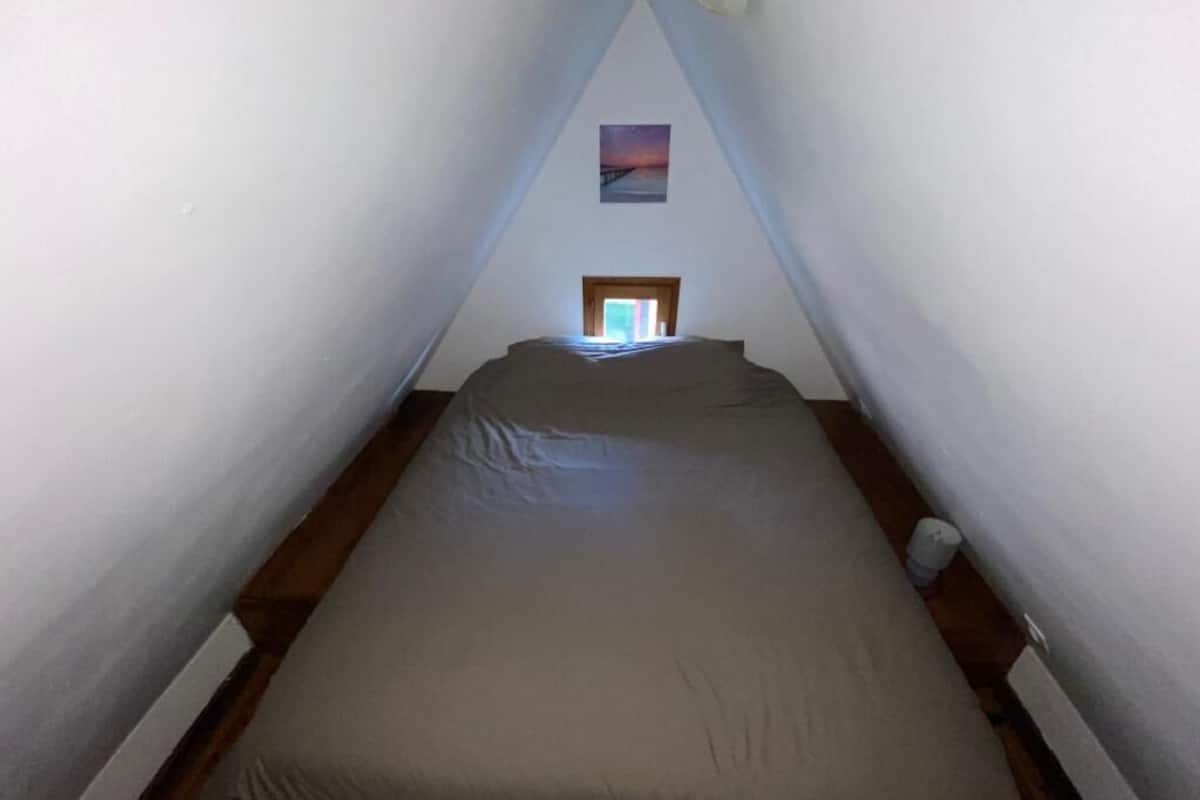 A narrow, triangular attic bedroom with a bed, a small window, and a picture on the wall.
