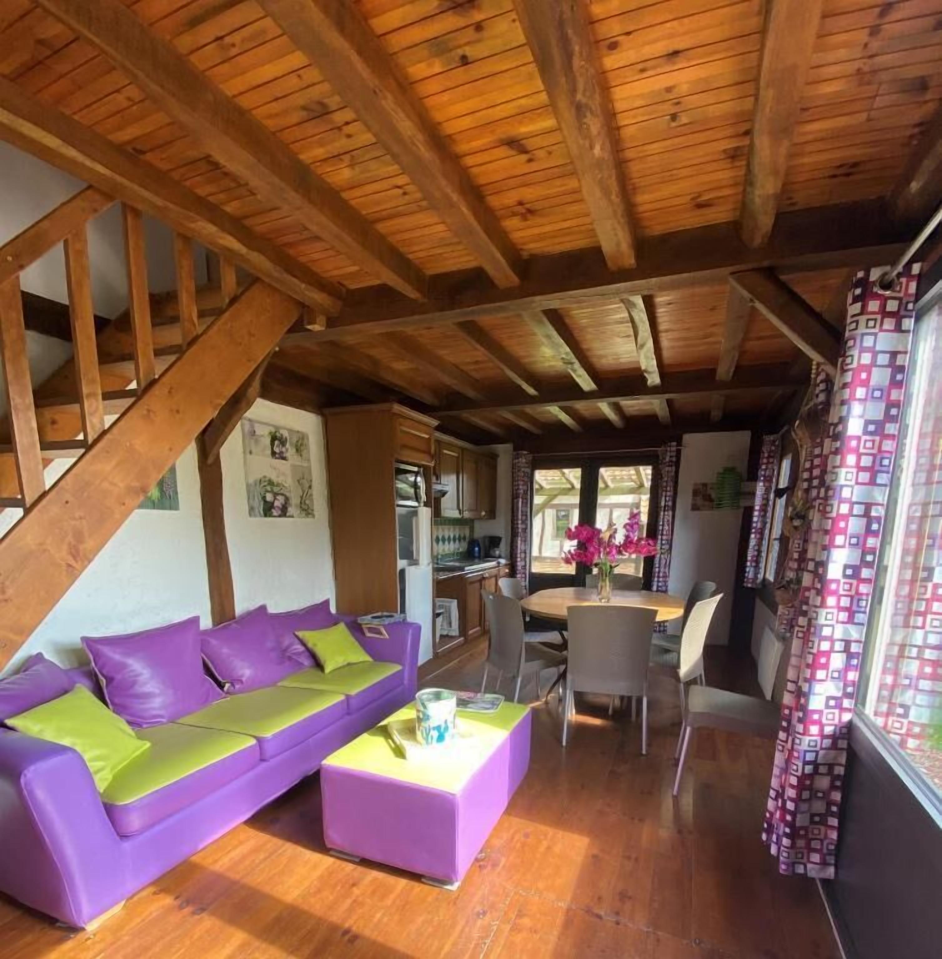 A cozy living room with a wooden ceiling, a purple sofa, a coffee table, and a dining area with chairs and a table.