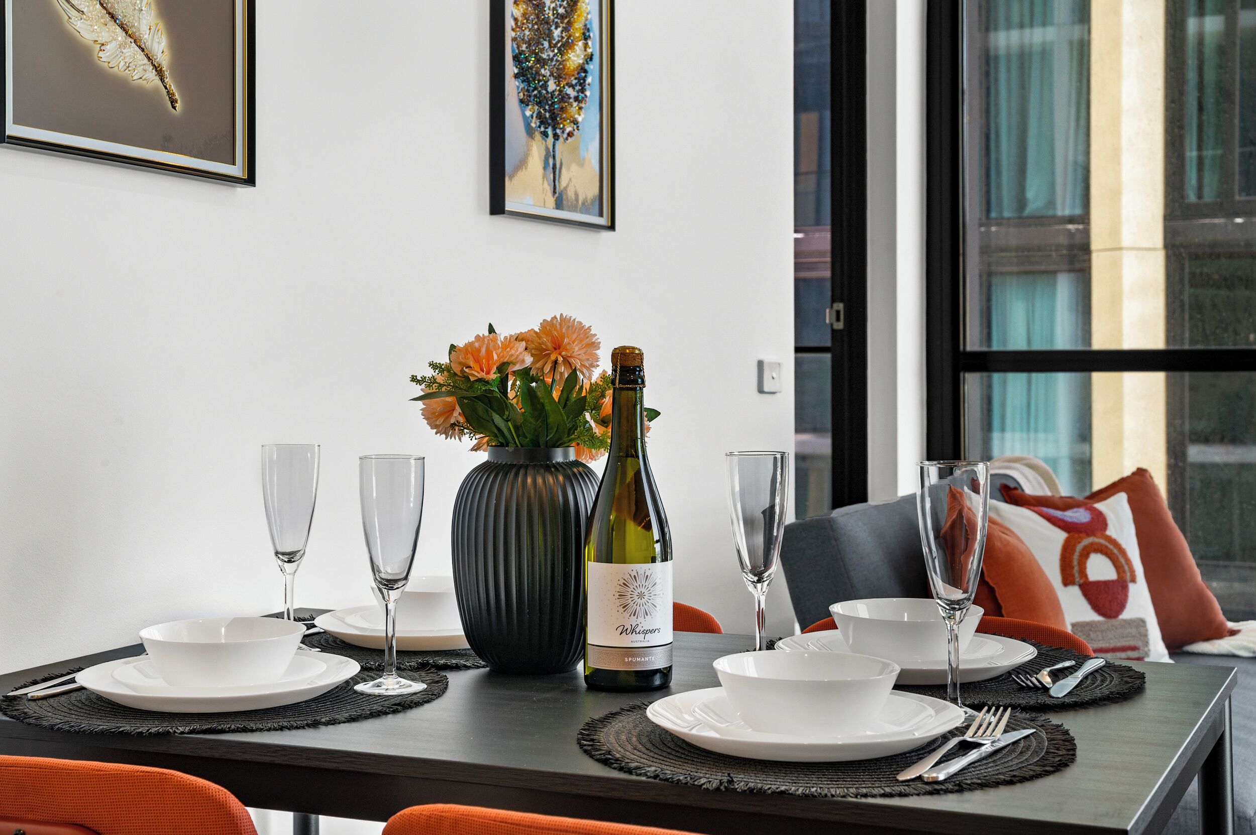 Une table à manger dressée avec de l’argenterie blanche, des couverts et des verres à vin, une bouteille de vin et un vase rempli de fleurs.