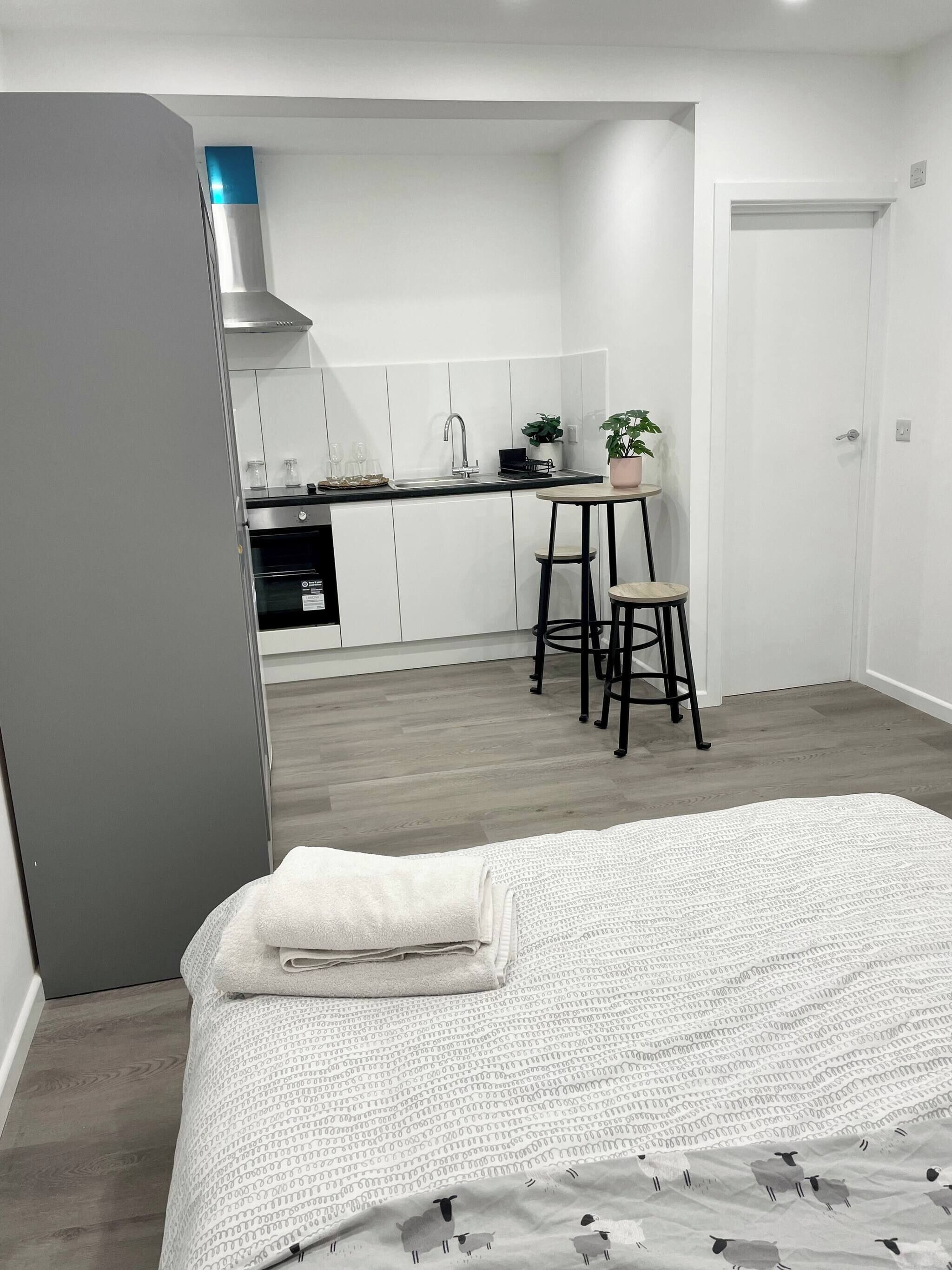 A compact bedroom with a bed, a small table, and a chair. Adjacent to the room is a kitchenette with a stove, sink, and cabinets.