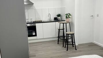 Une chambre compacte comprenant un lit, une petite table et une chaise. À côté de la chambre se trouve une kitchenette équipée d’une cuisinière, d’un évier et de placards.