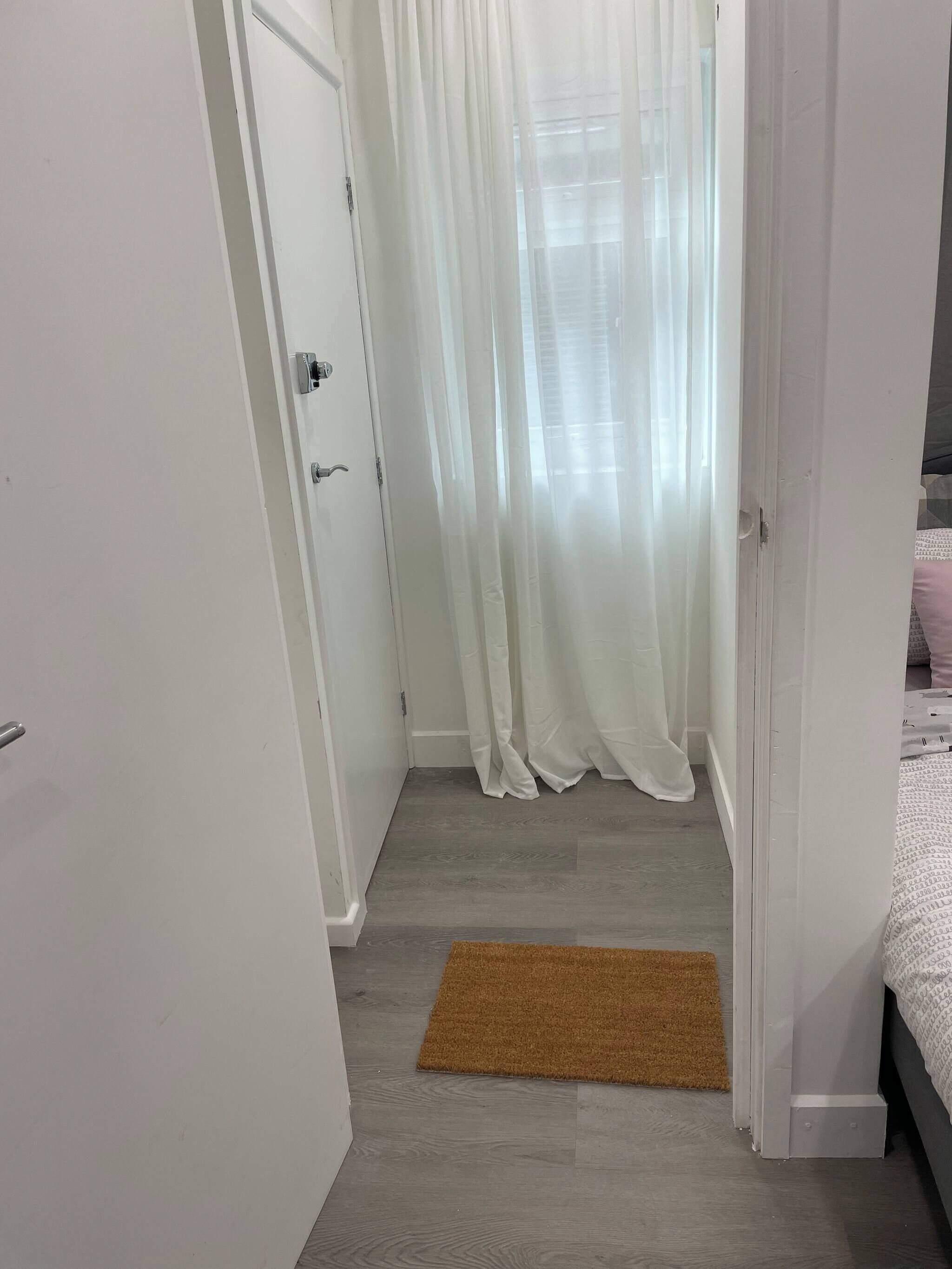 Een smalle gang met een houten vloer, een bruin tapijt, een witte deur en een bed met een beddegoed met een patroon.