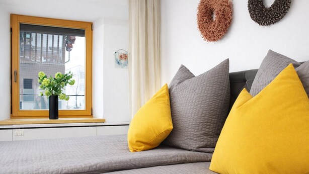 A modern bedroom with a grey sofa bed, two yellow pillows, a window with a view, and decorative wall art. - Dengg down town studios Apartment (Innsbruck)