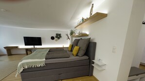Una camera da letto moderna con una comoda divano letto grigio, un tappeto a righe, una TV a parete e uno scaffale con oggetti decorativi.