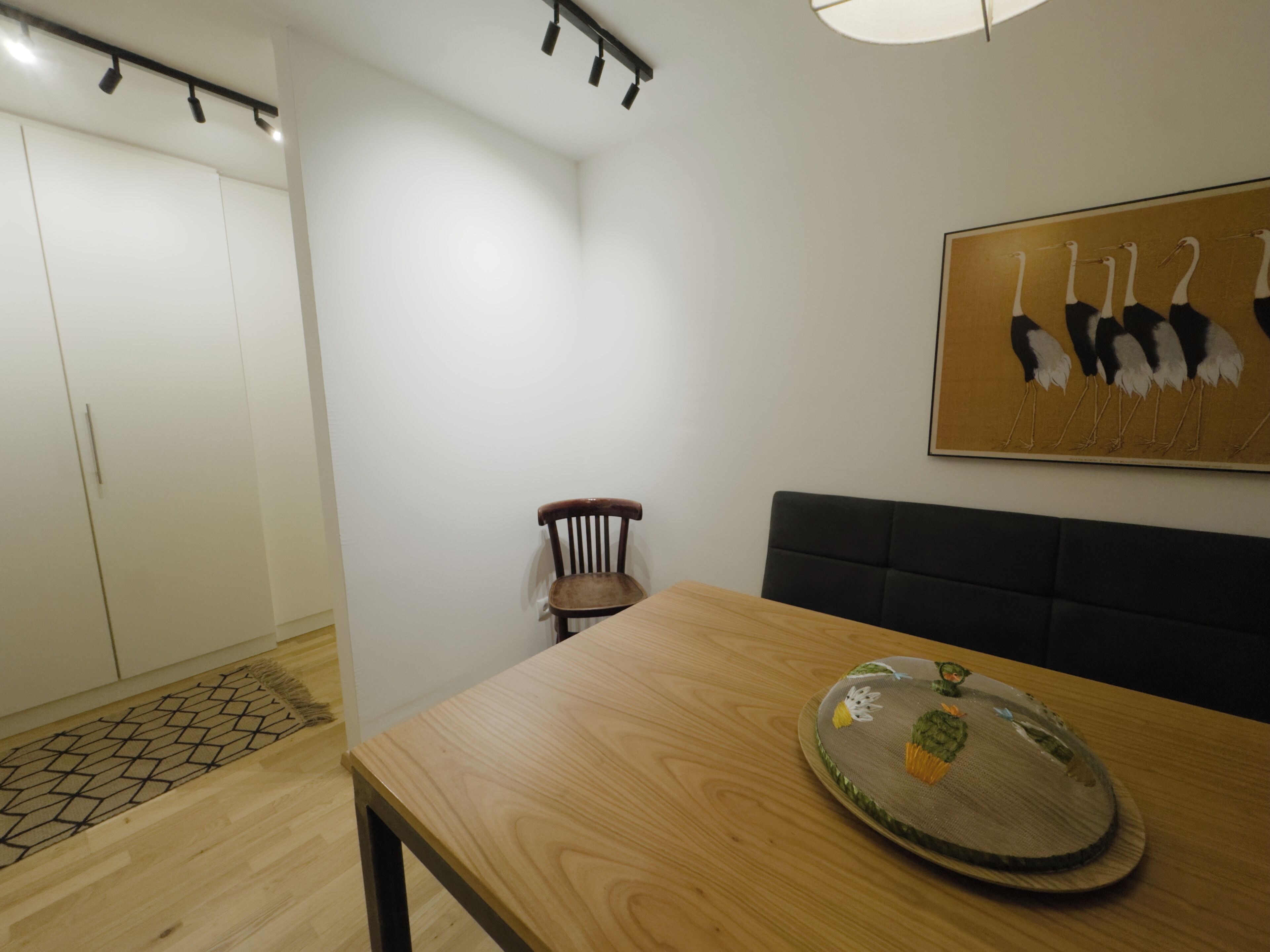 A room with a wooden table, a chair, a sofa, a wall-mounted artwork of birds, and a wardrobe.