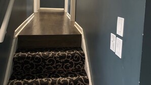 Uma escada estreita com carpete estampado, corrimão à esquerda e interruptor de luz na parede.