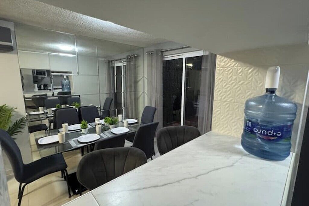A modern dining area with a large water dispenser, a kitchen in the background, and a view through a glass door.