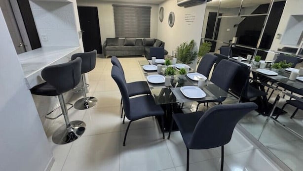 A modern dining area with a central table set for four, surrounded by black chairs. A grey sofa is against the wall, and a small plant is on the table.