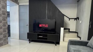 A modern living room with a flat-screen TV on a black stand, a grey sofa, and a staircase with a black railing.