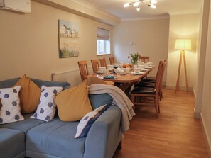 A dining area with a long table set for four, surrounded by wooden chairs. A sofa with patterned cushions is positioned in front of the table. - Cliff Cottage (Norwich)