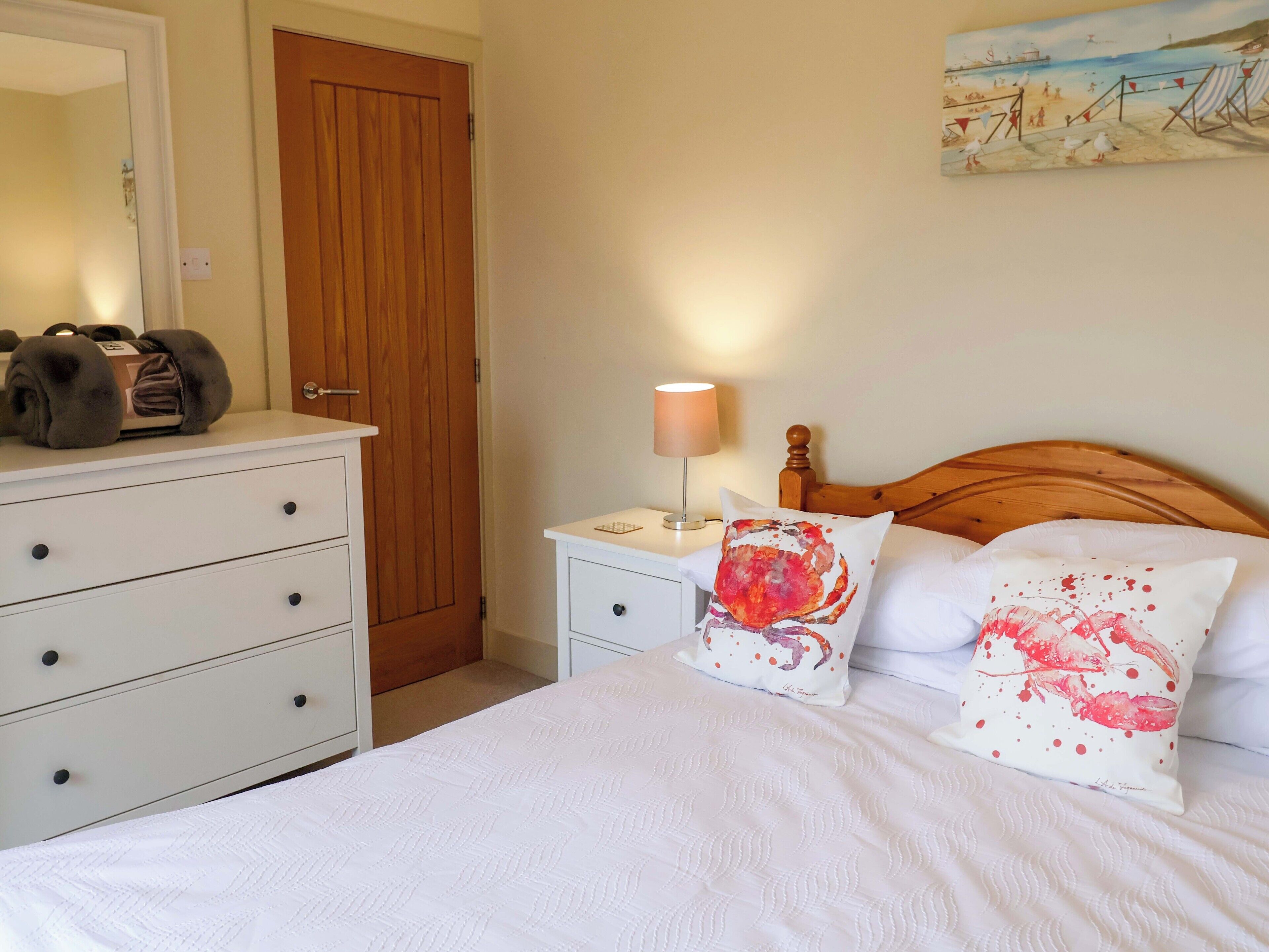 A bedroom with a wooden headboard, a white bed with two decorative pillows, a bedside table with a lamp, and a dresser with a mirror.