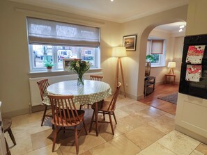 A dining area with a round table, chairs, a vase of flowers, a lamp, and a window with blinds. - Cliff Cottage (Norwich)