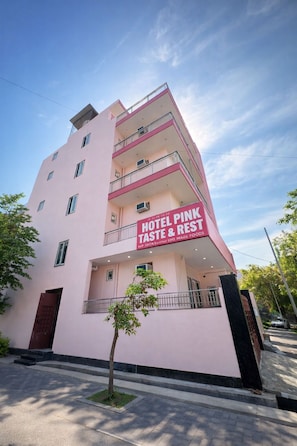 A pink multi-story hotel with balconies, a sign reading 'HOTEL PINK TASTE & REST,' and a tree in front. - Hotel Pink Taste & Rest  (Noida)