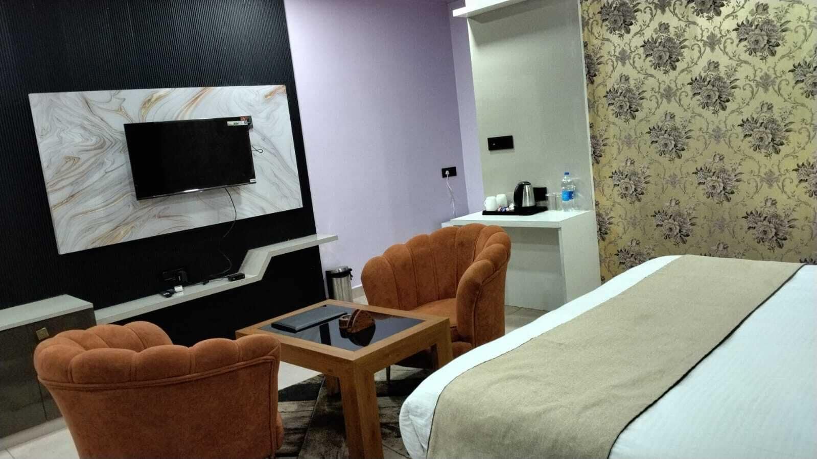 Quarto de hotel com cama, duas poltronas, uma mesinha, televisão fixada numa parede de mármore, escrivaninha com cadeira e uma garrafa de água sobre a escrivaninha.