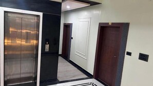 A hallway with a modern elevator, a door with a number plate, and a wall-mounted intercom. - Hotel Rashmi Grand (Khagaria)