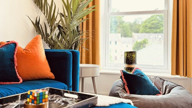 A modern living room with a blue sofa, a small table with a Rubik's cube, a potted plant, and a view of a cityscape through a large window. - The Ravens Inn, 4 - Bed, Sleeps 6, Long Term Rental Available (Ulverston)