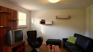 A living room with a television on a wooden stand, a black chair, a black sofa, a wooden coffee table, and a small table with a vase of flowers. - Himmelsbach Apartment with Terrace and Wi-Fi (Gengenbach)