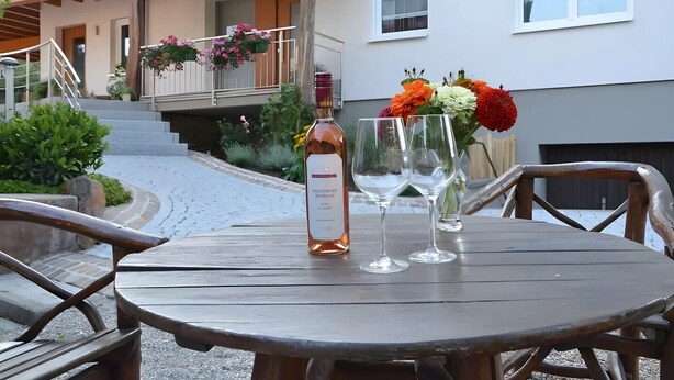 A wooden table with a bottle and two glasses, a balcony with hanging flower baskets, and a building with a white exterior. - Himmelsbach Apartment with Terrace and Wi-Fi (Gengenbach)