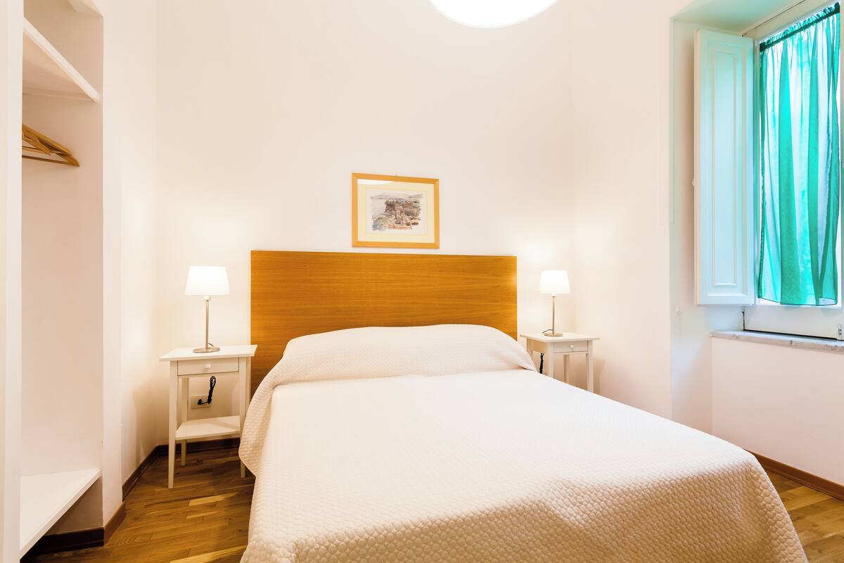 A neatly made bed with a wooden headboard, two bedside tables with lamps, a framed picture on the wall, and a window with curtains.