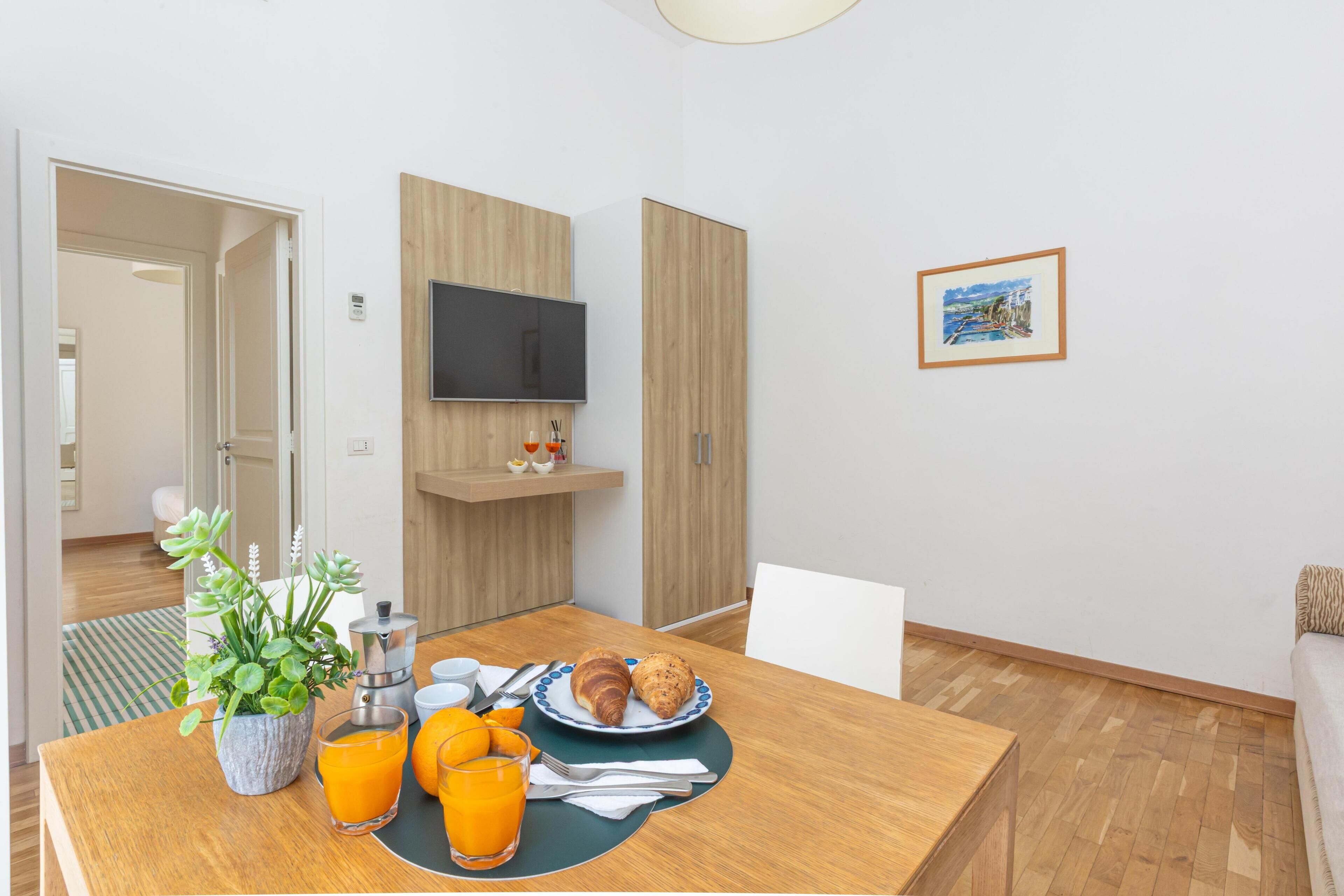 A modern room with a wooden dining table, breakfast setup, a TV mounted on the wall, and a framed picture.