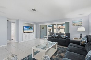 A modern living room with a flat-screen TV, a sofa, a coffee table, and a view of the outdoors through large windows. - Villa 13 Island House (Sarasota)