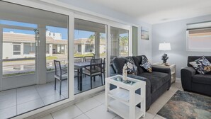 A modern living room with a glass sliding door leading to a balcony, a dining area with a table and chairs, and a grey sofa with floral cushions. - Villa 13 Island House (Sarasota)