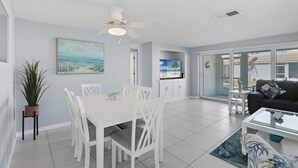 A modern living room with a white dining table, white chairs, a black sofa, and a glass coffee table. There is a ceiling fan, a painting on the wall, and a plant in a wicker basket. - Villa 13 Island House (Sarasota)