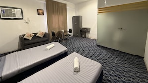 Habitación de hotel con dos camas individuales, una pequeña mesa con sillas, un sofá, un escritorio con silla, un televisor y un ventanal con cortinas.