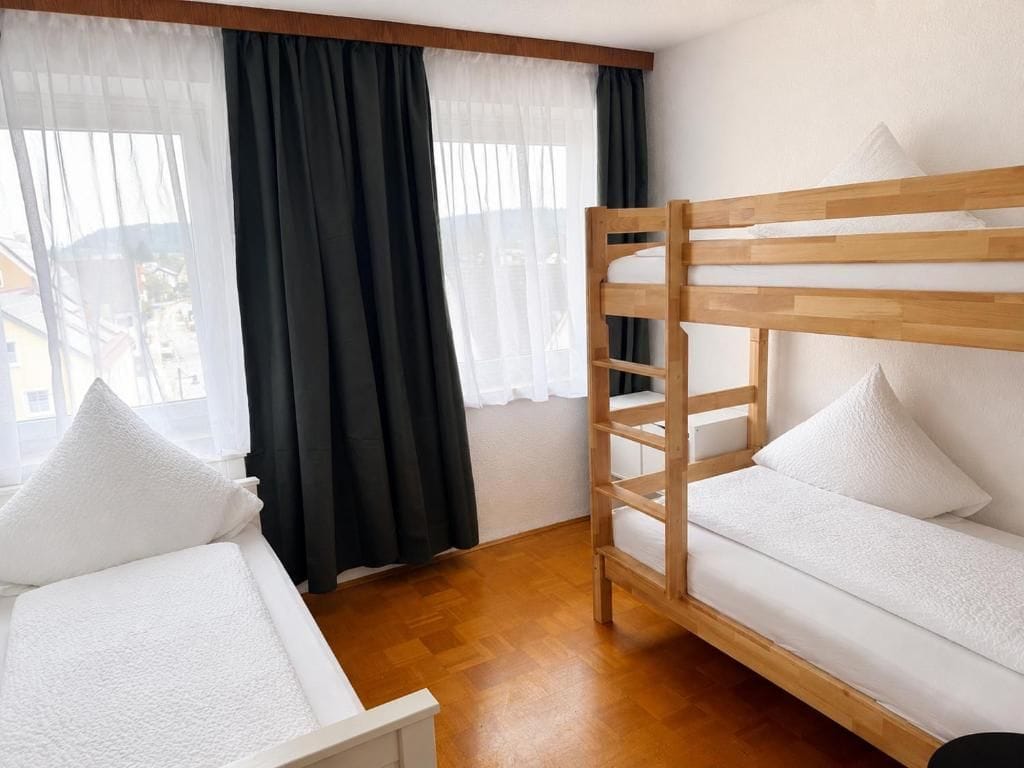 Una habitación con literas, una cama individual y un ventanal con cortinas traslúcidas.
