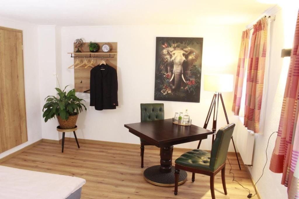 A room with a wooden floor, a dining table with chairs, a potted plant, a wall-mounted shelf with clothes and a clock, and a painting of an elephant.