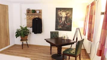 A room with a wooden floor, a dining table with chairs, a potted plant, a wall-mounted shelf with clothes and a clock, and a painting of an elephant.
