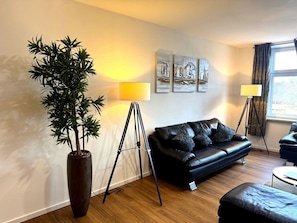 A modern living room with a black leather sofa, a large potted plant, floor lamps, and abstract wall art. - Gästehaus Jekle – Zimmer & Ferienwohnungen (Neuburg an der Kammel)