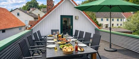 A rooftop dining area with a table set for six, surrounded by chairs, and an umbrella providing shade.