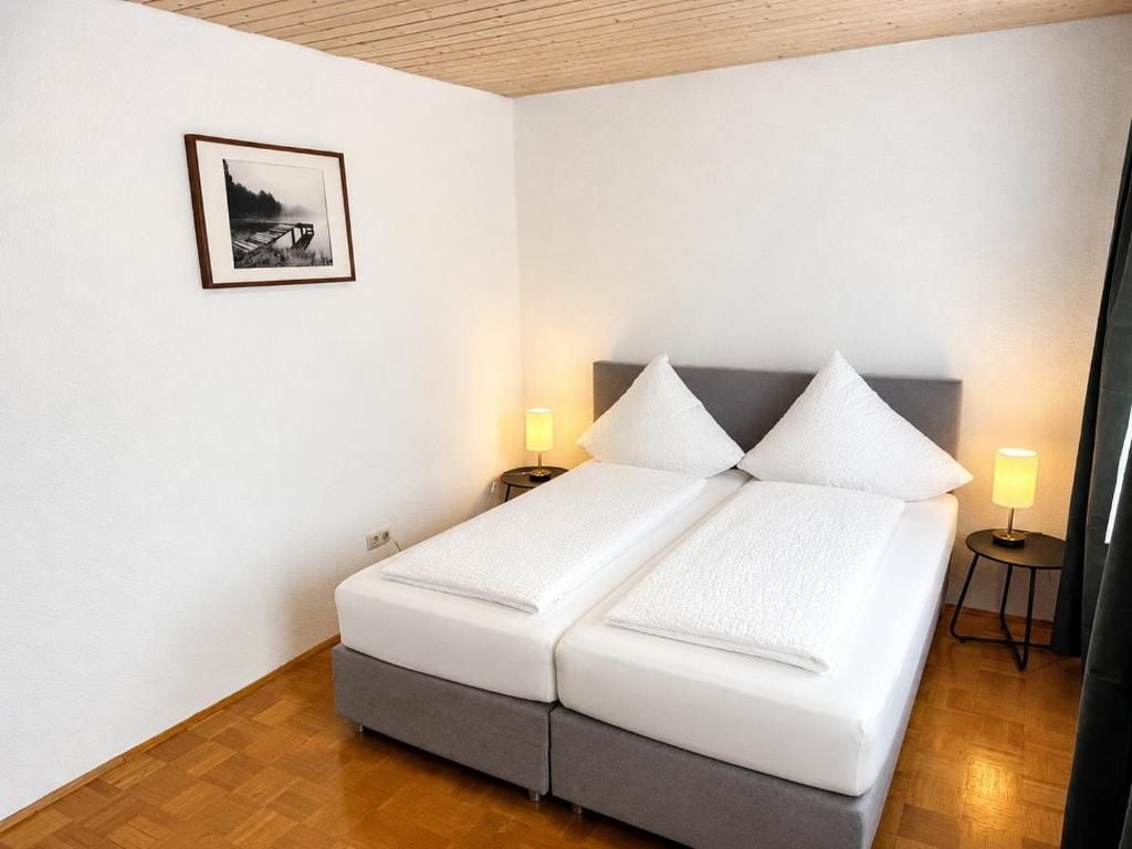Een tweepersoonsbed met een grijs hoofdbord, twee nachtkastjes met lampen en een ingelijst beeld aan de muur.