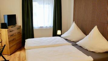 A hotel room with a bed, a TV on a wooden dresser, a window with curtains, and a bedside lamp.