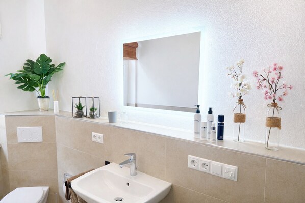 Un baño con lavamanos blanco, un espejo y artículos de tocador sobre la encimera.