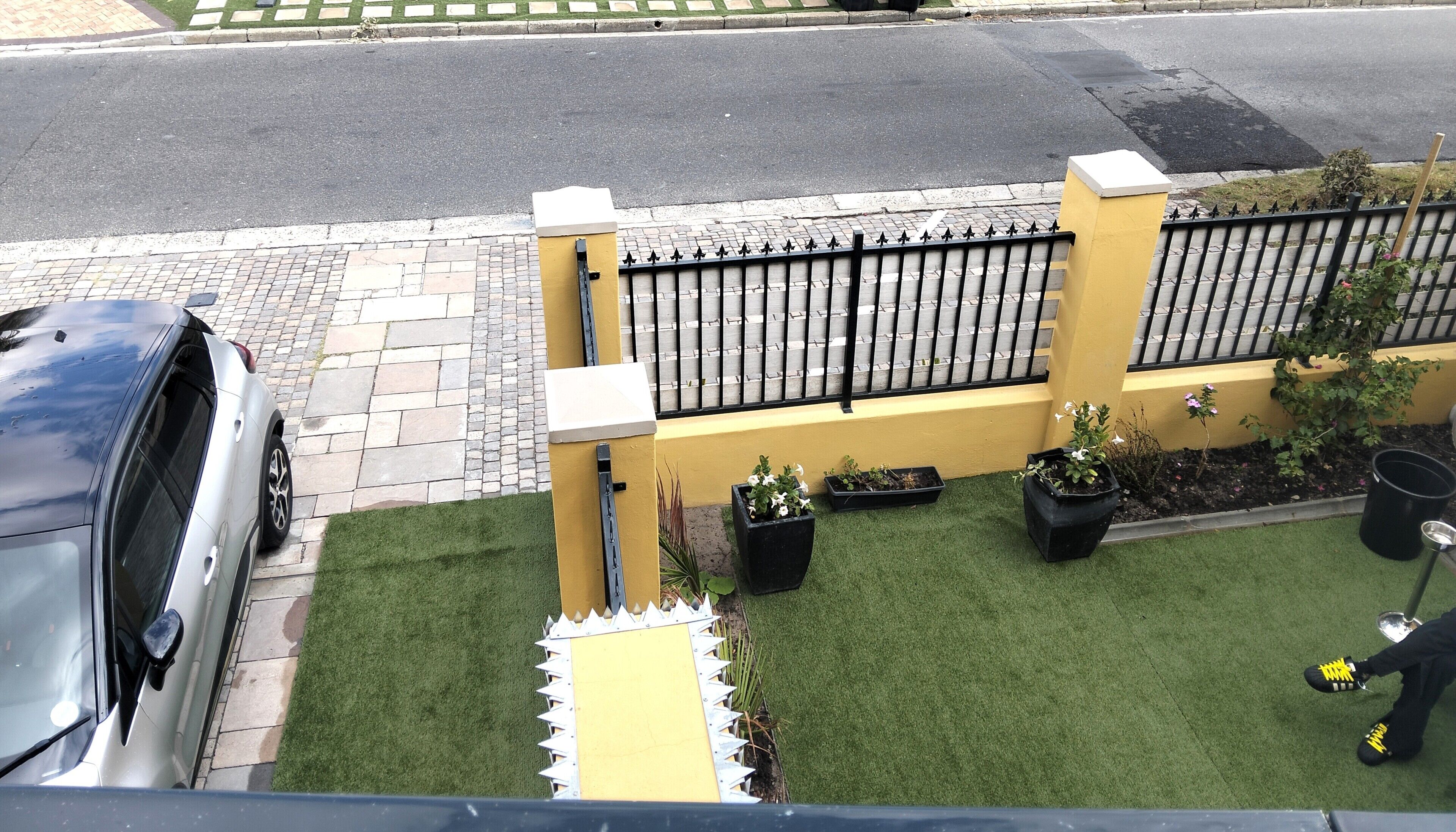 A residential area with a paved driveway, a yellow wall, a black metal fence, and potted plants.