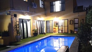 Une piscine dont le bord est revêtu de carreaux, entourée d’un patio agrémenté de plantes en pot et d’un espace salon abrité.