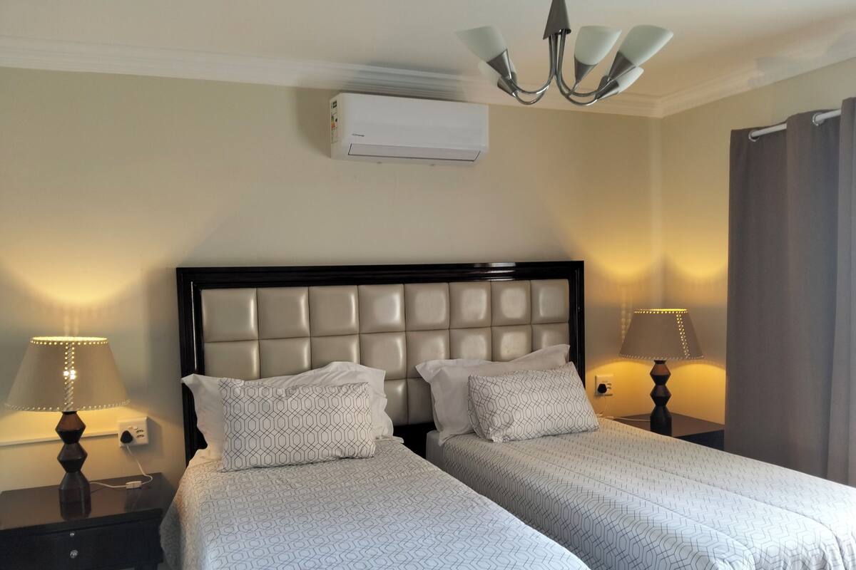 Habitación de hotel con cama doble, dos mesitas de noche con lámparas, unidad de aire acondicionado y candelabro.