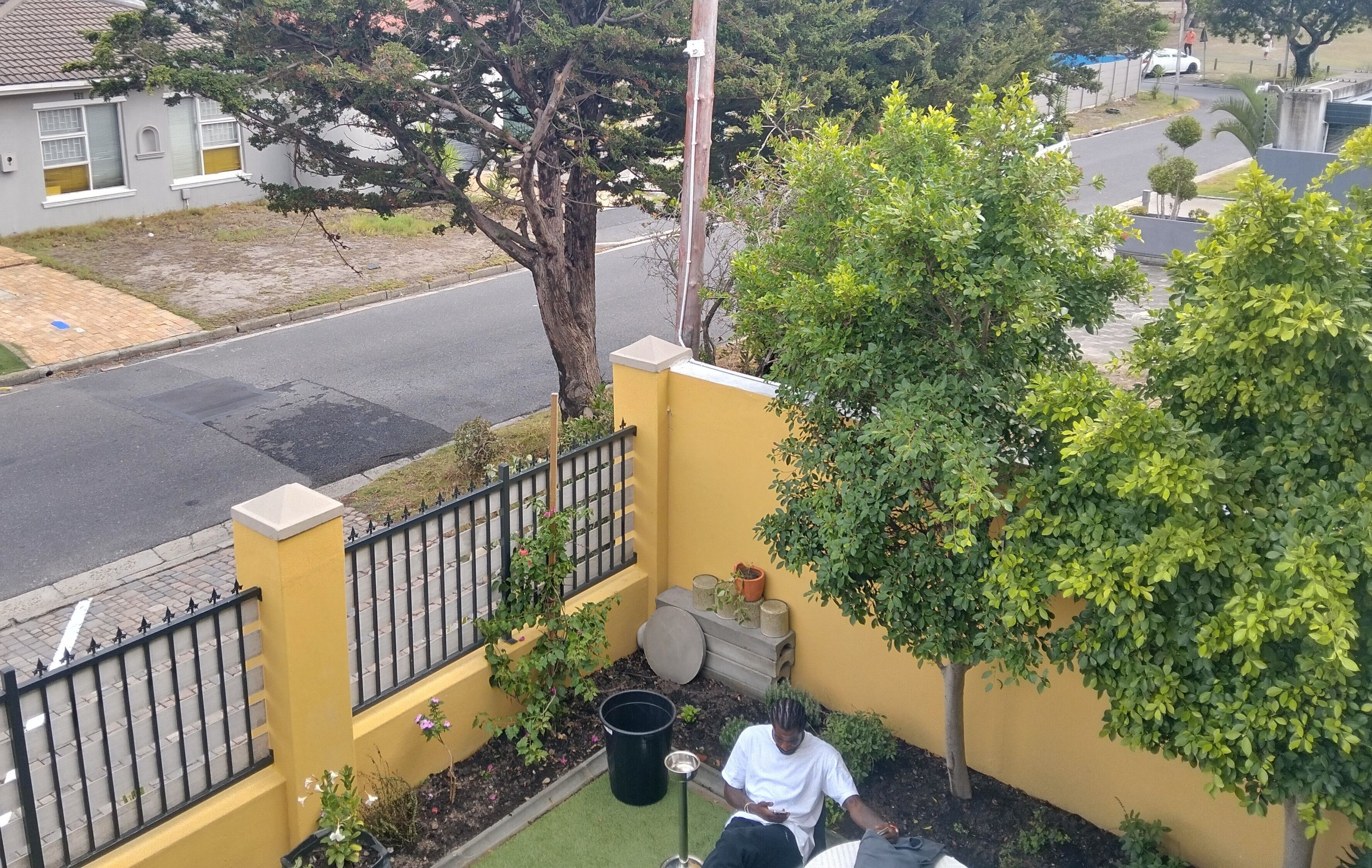 A residential area with a yellow wall, a black metal fence, a tree, and a person sitting on a bench.