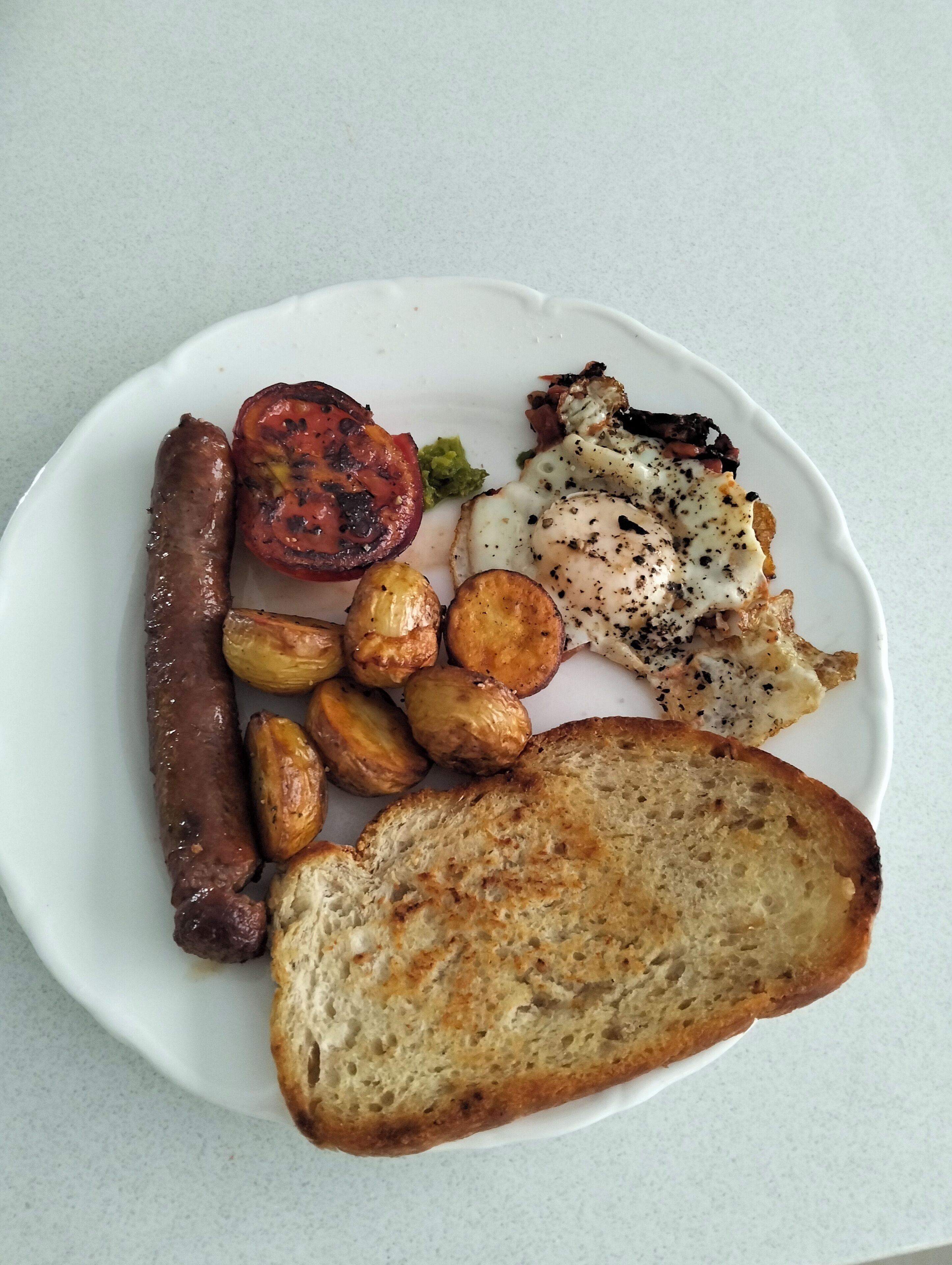A plate with a sausage, roasted potatoes, grilled tomato, fried egg, and a slice of toast.